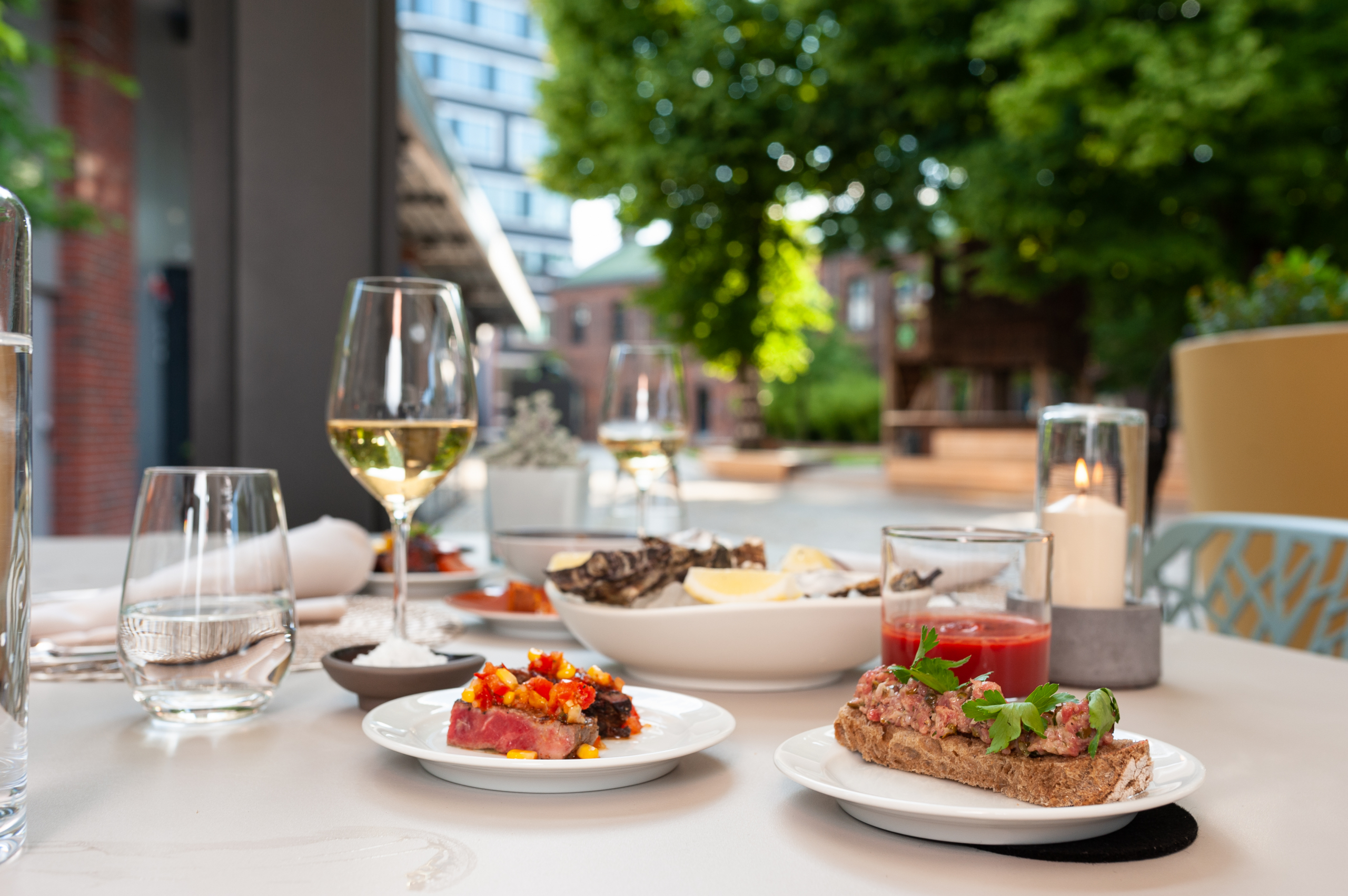 Ein sommerlicher Tisch, im Vordergrund kleine Speisen auf Tellern, Gläser mit Wein und Wasser, die Sonne scheint
