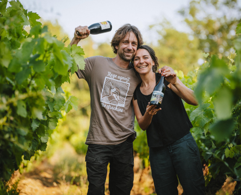 Flo Busch und seine Ehefrau Paola Ponisch halten jeweils eine Flasche Wein in der Hand, sie stehen im Grünen zwischen Weinreben